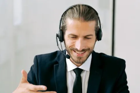 Smiling man in suit with headset working on laptop and gesturing during video call in office