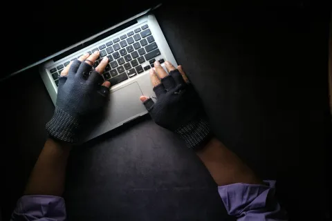 Hands in fingerless gloves typing on a laptop keyboard on a dark surface with sleeves rolled up.