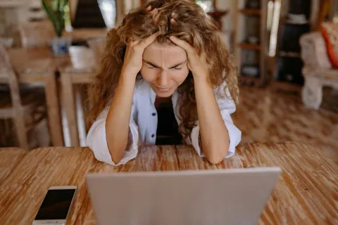 Frustrated woman sitting at wooden table holding head with hands in front of laptop and smartphone.