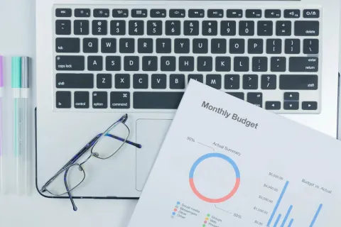 Glasses, pastel highlighters, and a monthly budget report on a laptop keyboard workspace.