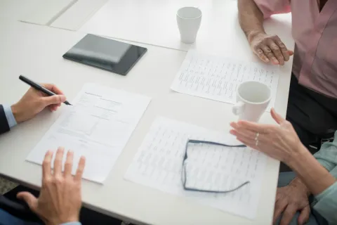 Two people reviewing and signing documents at a white table with coffee cups and eyeglasses.