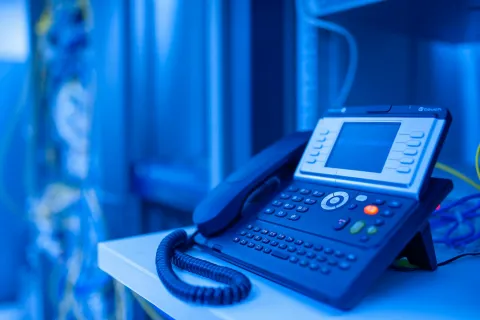 Black office desk phone with a digital screen and keypad on a white surface with blue background.