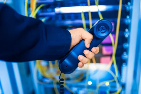 Person holding a blue telephone handset with network cables and server hardware in the background.