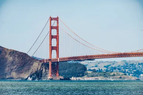 Golden Gate Bridge spans across blue water with rocky hills and clear sky in the background under daylight.