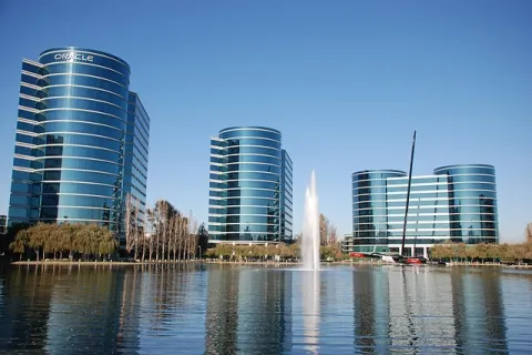 Modern office buildings with reflective glass facades beside a lake with a central fountain under a clear blue sky
