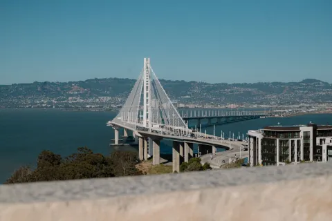 View of a modern cable-stayed bridge extending over a bay with city hills and buildings in the background.