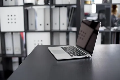 Open laptop on black office desk with blurred shelves and binders in the background