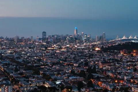 Evening aerial view of San Francisco cityscape with illuminated skyscrapers and Bay Bridge under a clear sky.