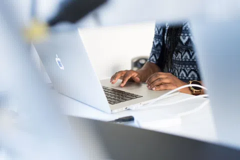 Person typing on a silver laptop with a sticker on the back in a bright office setting.