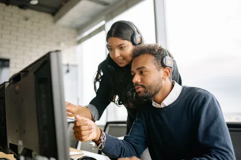 Two colleagues wearing headsets collaborate while looking at a computer monitor in a modern office.