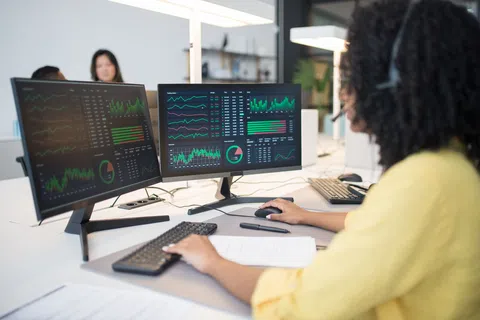 Woman wearing headset analyzes stock market data on dual monitors in a modern office environment.