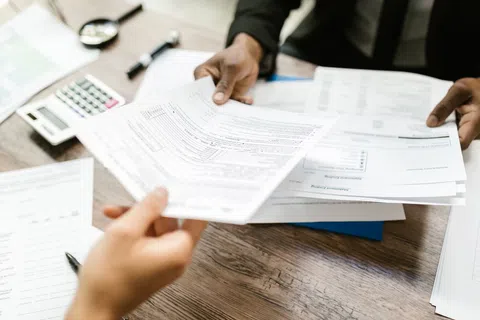 Two people exchanging and reviewing financial documents and forms at a wooden table with calculator and pen.