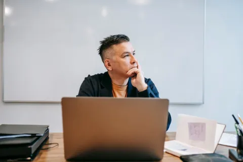 Man in casual office attire sitting at desk with laptop, pens, and papers, looking thoughtfully to the side.