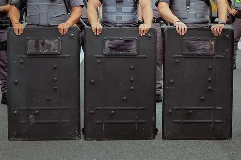 Three police officers holding large black riot shields standing side by side in uniform.
