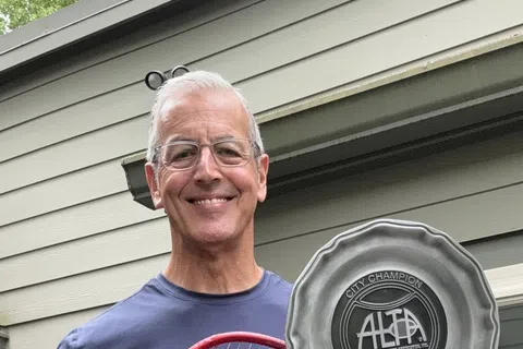 Smiling man holding a tennis racket and a City Champion ALTA 2026 trophy plate outdoors by a house.