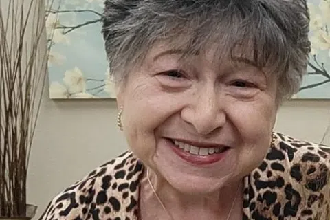 Smiling elderly woman wearing a leopard print jacket and heart key necklace in a floral background setting.