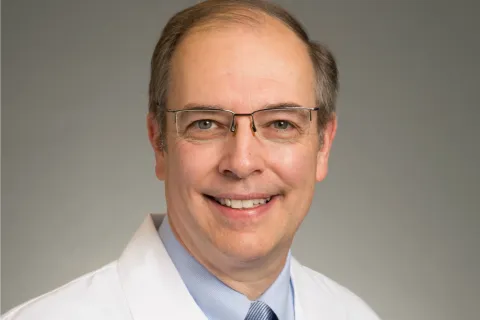 Smiling middle-aged male doctor wearing glasses, white coat, blue shirt, and tie against gray background
