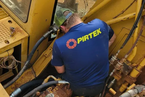 Worker in blue PIRTEK shirt repairs hydraulic machinery on yellow heavy equipment with various hoses and metal parts.