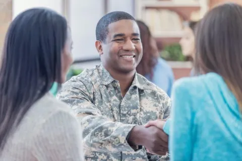 Smiling soldier in camouflage shaking hands with a woman in a friendly indoor setting.