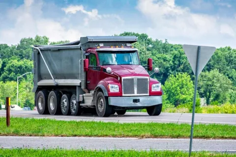 Red dump truck driving on rural road with green trees and blue sky background