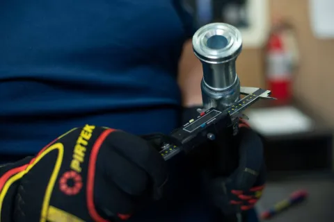 Worker wearing gloves measures metal pipe diameter with digital caliper in workshop setting.