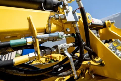 Close-up of yellow heavy machinery hydraulic system with metal pipes, valves, and hoses under bright daylight.