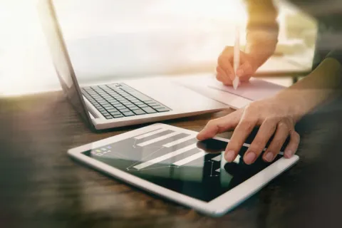 Person analyzing financial data on tablet with bar charts next to laptop and writing notes on paper