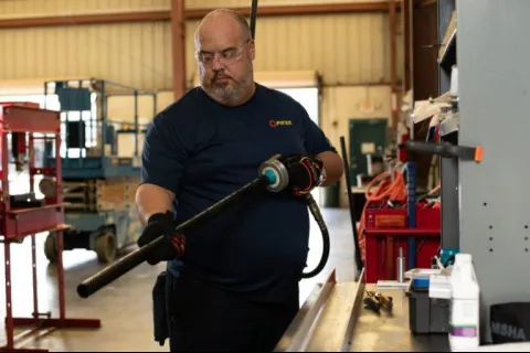 Man in safety glasses and gloves using a power tool on a metal pipe inside an industrial workshop.
