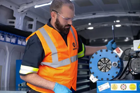 Technician wearing orange safety vest and gloves operating blue hydraulic equipment inside a van workshop.