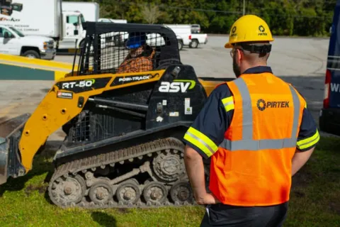 Construction worker in a Pirtek safety vest and helmet observing a yellow ASV Posi-Track RT-50 skid steer loader.
