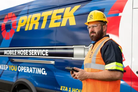 Worker in safety gear standing by a blue Pirtek mobile hose service van holding a device, ready for service.