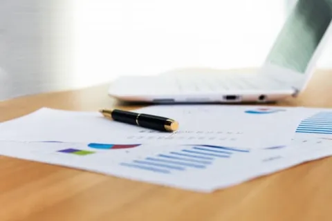 Black pen on financial charts and graphs with laptop on a wooden desk in a bright office.