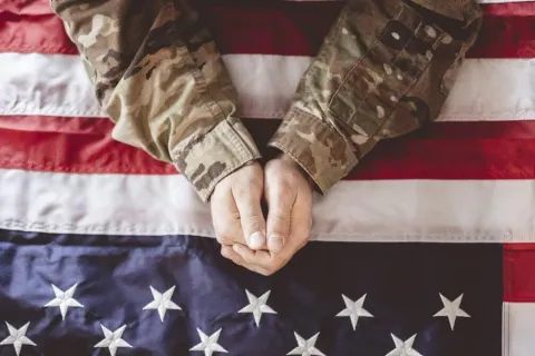 Hands of a soldier in camouflage uniform resting on a folded American flag.