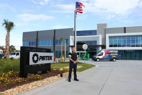 Man in uniform standing outside modern Pirtek building with company van and American flag visible.