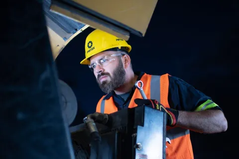 Worker wearing safety gear inspects machinery using a wrench under dim lighting conditions.