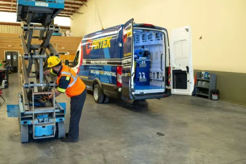 Technician in safety gear working on lift equipment next to a service van inside a spacious workshop.