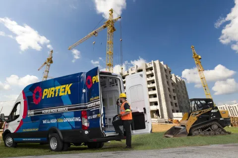Construction worker with safety gear accessing tools from a PIRTEK mobile hose service van at a building site with cranes.