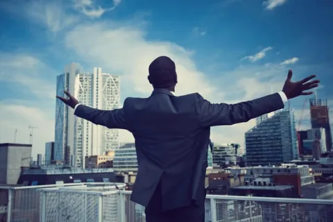 Businessman in suit stands on rooftop with arms outstretched overlooking city skyline under blue sky.