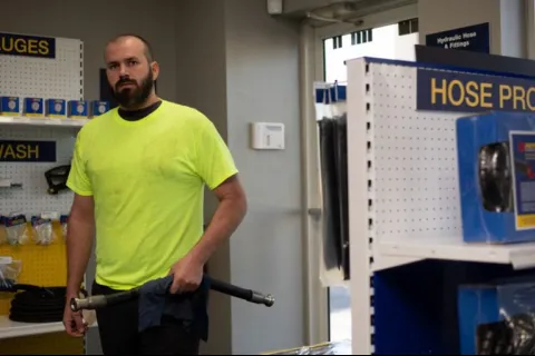 Man in a neon yellow shirt holding a hose in a hardware store aisle with hose products on shelves.