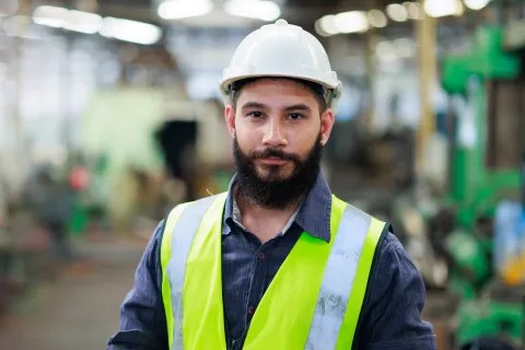 Confident male engineer in hard hat and reflective vest standing inside a factory or industrial setting.