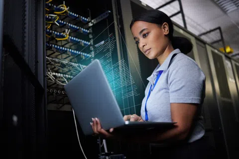 IT technician working on laptop in data center with server racks and digital code overlay in background