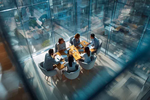 Aerial view of six professionals collaborating around a wooden table in a glass office with cityscape reflections.