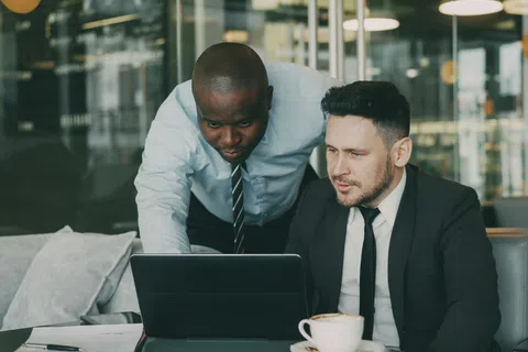 Two businessmen in office attire collaborating over a laptop with a coffee cup on the table nearby
