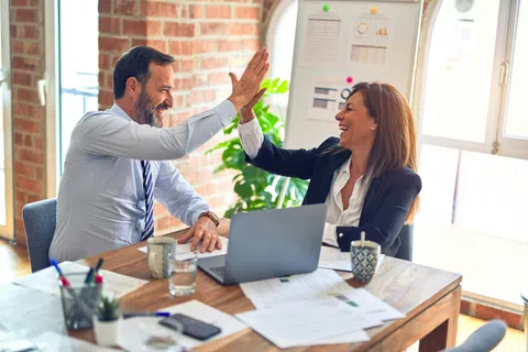 Two professional colleagues sharing a high five over a desk with laptop and paperwork in a bright office