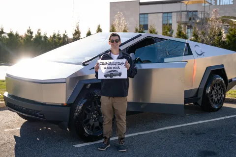 Man standing beside a silver Tesla Cybertruck holding a congratulatory sign in a parking lot on a sunny day