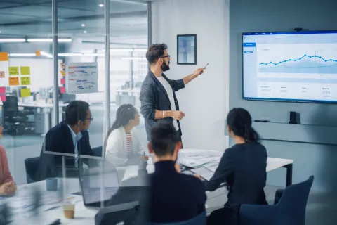 Business team in meeting room watching man present data analytics on large screen with graphs and charts