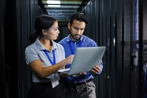 Two IT professionals examining data on a laptop inside a modern data center server room.