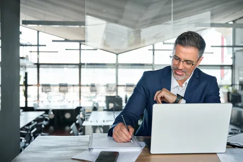 Businessman in a blue suit working on laptop while taking notes in a bright modern office.