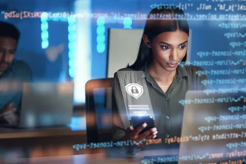 Woman focused on smartphone and laptop with digital security icons and code overlay in modern office environment