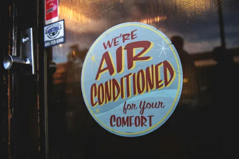Window sign stating We're Air Conditioned for Your Comfort on a glass door with reflections in warm light